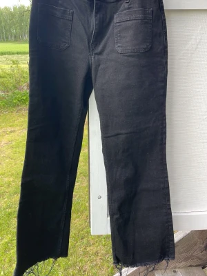 Svarta raka jeans med rå kant - Säljer ett par svarta jeans med raka ben och råa, fransiga benslut. Jeansen har hög midja och stora fickor både fram och bak. Perfekta för en avslappnad och trendig look.