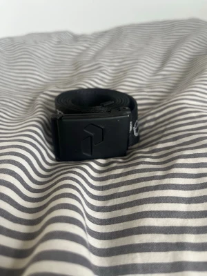 Svart Peak Performance bälte - Snyggt svart bälte från Peak Performance med justerbart spänne i metall och logga tryckt längs hela bandet. Perfekt accessoar för att lyfta din outfit med en sportig touch. Klassisk design som funkar till det mesta.