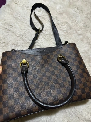 Fint handväska - Snygg handväska i klassiskt brunt damier-mönster. Väskan har både korta handtag och en längre axelrem i mörkbrunt läder samt guldfärgade metalldetaljer. Perfekt storlek för det viktigaste och ger en lyxig touch till din outfit.