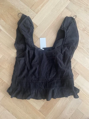 Svart blus med volang och spetsdetaljer - Snygg svart blus med volangkant nedtill och spetsdetaljer längs axlar och framtill. Blusen har en luftig passform och är ärmlös med breda axelband. Perfekt att styla med jeans eller kjol för en trendig look. Originellt pris: 299kr