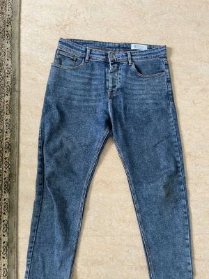 Blå slim fit jeans från Blue Hand - Säljer ett par klassiska blå slim fit jeans från Blue Hand. Jeansen har fem fickor, bälteshällor och stängs med knapp och dragkedja. Materialet är denim i bomull och färgen är en tvättad mellanblå nyans med subtila slitningar vid låren. Dessa passar som slim fit