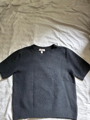 Svart stickad tröja från H&M - En enkel och stilren svart stickad tröja från H&M med korta ärmar och rund hals. Tröjan har ribbade muddar vid hals och nederkant och är gjord i ett mjukt stickat material. Perfekt att matcha med jeans eller kjol.