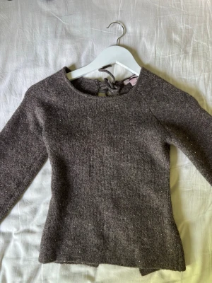 Brun stickad tröja med knytning bak - Mysig brunmelerad stickad tröja med långa ärmar och unik knytning i ryggen. Tröjan har en rund halsringning och är figurnära i modellen. Perfekt för dig som gillar en enkel men ändå speciell look.