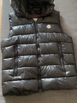 Svart dunväst från Moncler med huva - Snygg svart dunväst från Moncler med avtagbar huva och stora vita bokstäver på huvan. Västen har två dragkedjeförsedda fickor, quiltad design och Moncler-logga på bröstet. Tillverkad i glansigt material som ger en exklusiv look.
