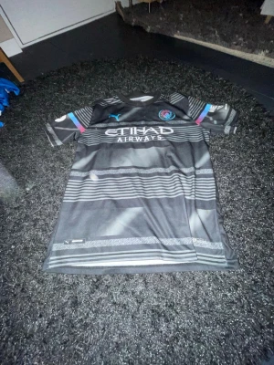 Manchester City Haaland matchtröja - Snygg svart Manchester City matchtröja från Puma med Haaland och nummer 9 på ryggen. Tröjan har mönstrade ränder, klubbmärke på bröstet och sponsortryck. Färgdetaljer i blått och rosa på ärmarna. Perfekt för fotbollsfans! Priset kan diskuteras 