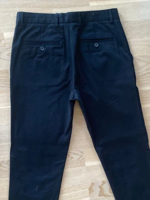 Svarta chinos med raka ben - Snygga svarta chinos med raka ben och klassisk passform. Byxorna har två bakfickor med knapp och hällor för bälte. Perfekta för en stilren look och tillverkade i mjukt bomullstyg.
