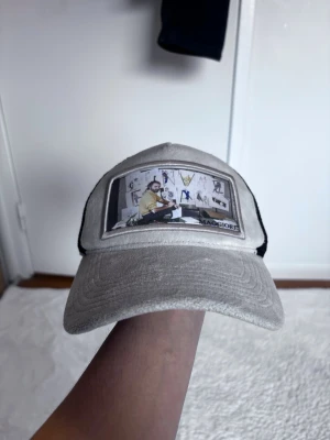 Grå keps med konstmotiv från Maggiore - Säljer en grå keps från Maggiore med svart mesh på sidorna och ett stort konstnärsmotiv framtill. Kepsen har böjd skärm och justerbar baksida. Perfekt för dig som gillar unik streetstyle och vill sticka ut med cool design.