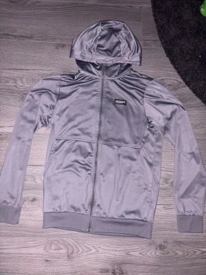 Grå hoodie från McKenzie - Snygg grå hoodie från McKenzie med dragkedja framtill och huva. Tillverkad i ett glansigt, mjukt material som känns sportigt. Har ribbade muddar vid ärmslut och nederkant samt en liten logotyp på bröstet. Perfekt för en chill och avslappnad stil.