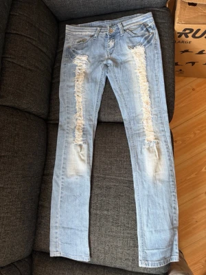 Tommy Hilfiger jeans  - Säljer ett par ljusblåa Tommy Hilfiger jeans, med slitningar på benen fram och märken och andra fina detaljer på baksidan, i mkt bra skick. 