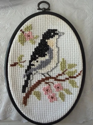 Broderad tavla med fågelmotiv - Säljer en oval broderad tavla med svart och vit fågel på en gren med rosa blommor och gröna blad. Tavlan har svart ram och broderiet är gjort på vit aidaväv. Perfekt som unik väggdekoration.
