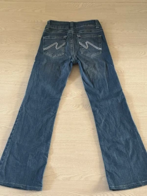Nelly Stich Jeans - Säljer ett par klassiska blå bootcut jeans från Wrangler med snygga kontrastsömmar och broderade bakfickor. Jeansen har normal midja och är tillverkade i slitstark denim. Perfekta för dig som gillar retrostil och vill ha ett par jeans med schysst passform.
