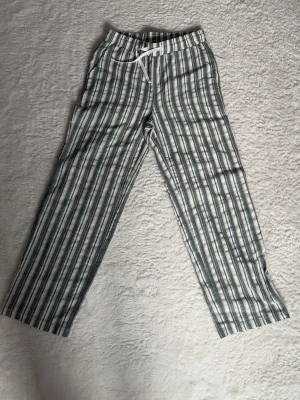 Randiga pyjamasbyxor med dragsko - Säljer ett par pyjamasbyxor med gröna och vita vertikala ränder. Byxorna har resår i midjan och vit dragsko för justerbar passform. Tillverkade i ett mjukt och lätt material som passar perfekt för att chilla hemma.