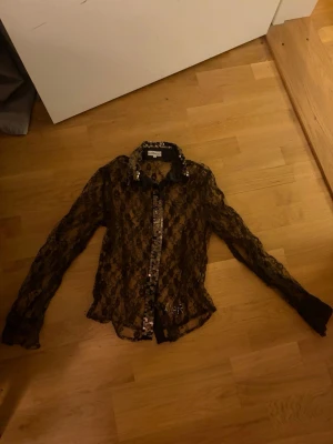 Svart spetsblus med paljettdetaljer - Säljer en transparent svart skjorta från Dolce Ribelle i spets med blommigt mönster. Skjortan har långa ärmar och är dekorerad med paljetter längs kragen och knäppningen. Perfekt för dig som gillar att sticka ut med detaljer och unik design.
