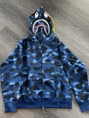 Blå camo hoodie med hajmotiv - Säljer en blå camo hoodie från BAPE med dragkedja och hajmotiv på huvan. Hoodien har känguruficka framtill och ribbade muddar. Färgglada detaljer på huvan och klassisk streetwear-stil. Perfekt för dig som gillar unika och iögonfallande plagg.