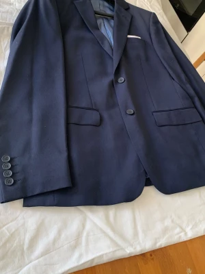 Mörkblå kavaj Dressmann Slim Fit - Stilren mörkblå kavaj från Dressmann i modellen Slim Fit. Kavajen har två knappar framtill, tre fickor och fyra knappar vid ärmsluten. Fodrad insida i ljusblått tyg och klassisk passform som ger en snygg siluett.