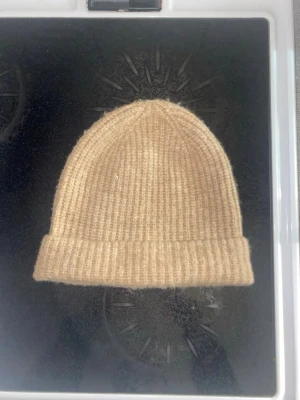 Beige ribbstickad mössa - Säljer en beige ribbstickad mössa med uppvikt kant. Mössan har en klassisk form och är stickad i ett mjukt material som känns skönt mot huden. Perfekt för att hålla värmen under kyliga dagar. inte kommit till användning 