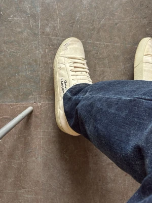 Saint Laurent vita låga sneakers - Säljer ett par vita låga sneakers från Saint Laurent med klassisk design och snörning. Skorna har textdetalj på sidan och är tillverkade i canvas med gummisula