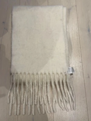 Vit ullhalsduk från H&M med fransar - Mysig vit halsduk från H&M i ullblandning med klassiska fransar i ändarna. Perfekt att svepa runt halsen för en stilren look. Materialet är mjukt och värmande, och färgen är lätt att matcha med olika outfits.