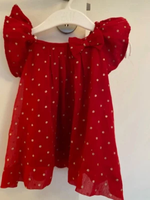 Röd prickig klänning med rosett - Söt röd kortklänning med vita prickar och stora puffärmar. Klänningen har en dekorativ rosett framtill och knappar längs ryggen. Perfekt för dig som gillar lekfulla detaljer och ett färgstarkt uttryck.