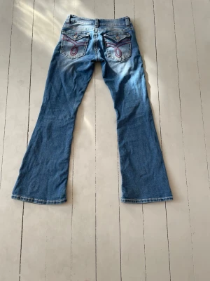Blå bootcut jeans med slitningar - Säljer ett par blå bootcut jeans med slitna detaljer på knäna och dekorativa sömmar på bakfickorna. Jeansen har klassisk femficksdesign och är tillverkade i denim med lätt stretch. Perfekta för en avslappnad och trendig look. Vi har sytt vid gylfen eftersom sömmen gick upp där vid andra användningen men syns inte alls och är väl sytt. Säljer då de är för små för mig. 