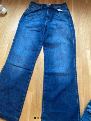 Blåa raka jeans med hög midja - Säljer ett par klassiska blå jeans med rak passform och hög midja. Jeansen har fem fickor, knappgylf och är tillverkade i ett robust denimtyg. Perfekta för en avslappnad och trendig look.