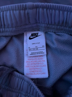 Grå Nike Dri-FIT mjukisbyxor XL - Säljer ett par grå mjukisbyxor från Nike i Dri-FIT material. Byxorna har resår i midjan och muddar vid bensluten samt en vit sidoremsa med Nike-loggor längs ena benet. Perfekta för chill eller träning. PRISET KAN DISKUTERAS!