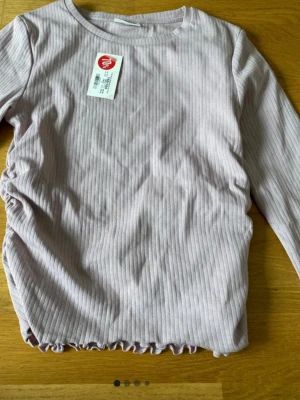 Ljuslila ribbad långärmad topp - Supersöt ljuslila ribbad topp från Kappahl med långa ärmar och rund halsringning. Toppen har en lätt rynkad detalj i sidan och vågig kant nertill för en extra touch. Perfekt att styla med jeans eller kjol för en chill look.