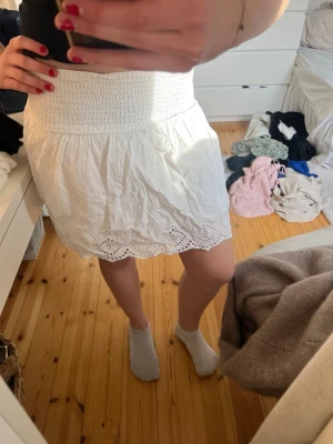 Vit kort kjol med broderade detaljer - Supersöt vit kort kjol med bred smock i midjan och broderade spetsdetaljer längs fållen. Kjolen har en luftig känsla och passar perfekt till varma dagar. Den har en feminin och fräsch look som är lätt att matcha med olika toppar.