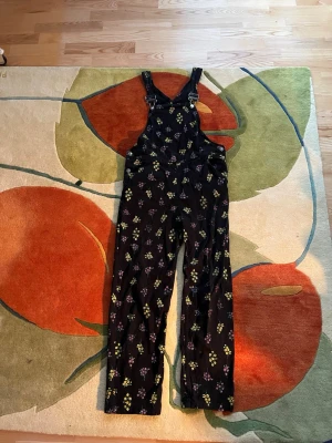 Svart blommig jumpsuit med hängslen - Snygg svart jumpsuit med raka ben och justerbara hängslen. Täckande blommönster i gult och lila. Väl använda men väl omhändertagna 