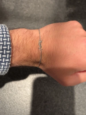Silverfärgat YSL-armband - Snyggt och stilrent armband i silverfärg från Yves Saint Laurent. Tunt kedjearmband med YSL-logga som detalj framtill. Perfekt accessoar för dig som gillar minimalistisk och exklusiv design. Justerbar längd med smidig låsning. Fint att ge även bort som present