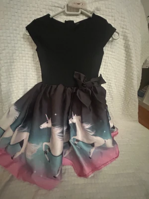 Svart klänning med enhörningsmotiv - Supersöt klänning med svart överdel och färgglad kjol med enhörningar i vitt och rosa mot en blå bakgrund. Kjolen har en stor rosett i midjan och klänningen har korta ärmar. Perfekt för dig som älskar magiska motiv! Vet ej storlek passar ungefär mellan 5-7 år