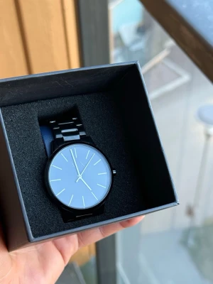 Svart minimalistisk klocka i metall - Stilren svart klocka från Axcent med metallarmband och minimalistisk urtavla. Klockan har tunna visare och indexmarkeringar i silver och guld, vilket ger en modern och clean look. Perfekt accessoar för dig som gillar enkel och tidlös design.