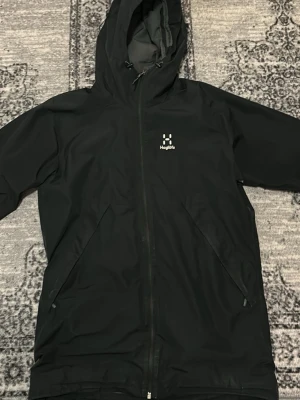 Svart vindjacka från Haglöfs med huva - Svart vindjacka från Haglöfs med dragkedja framtill, två snedställda fickor och justerbar huva. Jackan har minimalistisk design och logga på bröstet. Perfekt för blåsiga dagar och outdoor-aktiviteter.