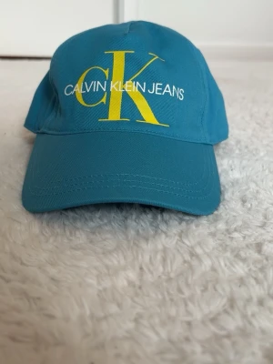 Turkos keps från Calvin Klein Jeans - Snygg turkos keps från Calvin Klein Jeans med stort gult CK-tryck och vit logotyp framtill. Klassisk böjd skärm och justerbar passform. Perfekt accessoar för en cool och avslappnad stil.