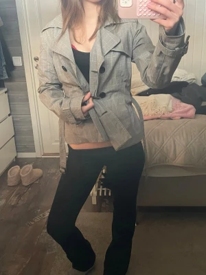 Grå rutig trenchcoat med bälte - Snygg grå trenchcoat med diskret rutmönster och svarta knappar. Jackan har klassisk krage, axelklaffar och tillhörande bälte i midjan för en markerad siluett. Perfekt för dig som gillar stilrena och trendiga ytterplagg.