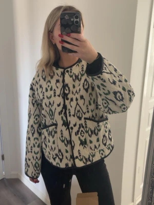 Mönstrad cardiganjacka i vitt och grönt - Säljer en oversized cardiganjacka med svart, grönt och vitt mönster. Jackan har svarta detaljer vid kanter och fickor samt två framfickor. Rund halsringning och knäppning framtill. Perfekt för dig som gillar statement-plagg.