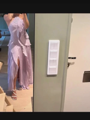 Ljuslila långklänning med volanger - Superfin ljuslila långklänning med volangdetaljer och plisserat tyg. Klänningen har en asymmetrisk axel och en slits framtill som ger en snygg touch. Perfekt för dig som vill sticka ut med en unik och elegant look.