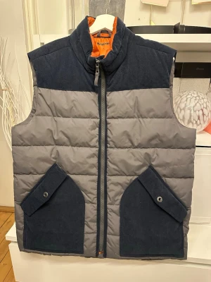 Grå och marinblå dunväst från Kappahl - Snygg dunväst från Kappahl i grått och marinblått med orange foder. Västen har två stora fickor framtill med lock och knappar samt hög krage och dragkedja. Perfekt för lager-på-lager och en cool streetstil.