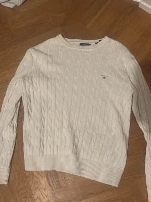 Vit kabelstickad tröja från GANT - Snygg vit kabelstickad tröja från GANT med rund hals och diskret logga på bröstet. Tröjan är tillverkad i mjukt bomullsmaterial och har ribbade muddar vid ärmslut och nederkant. Perfekt för dig som gillar klassisk och stilren design. Storlek: 158/164 cm, 13-14 år. 