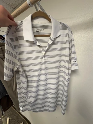 Vit randig polotopp från Nike Golf - Vit polotopp från Nike Golf med tunna gråa ränder och klassisk krage. Materialet är polyester och toppen har korta ärmar samt Nike-logga på ena ärmen. Perfekt för dig som gillar sportig stil och vill ha något bekvämt och snyggt.
