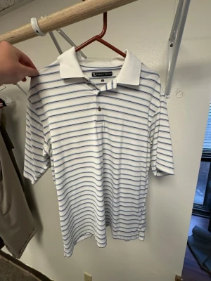 Vit randig pikétröja från Pebble Beach - Golfpiké. Vit pikétröja från Pebble Beach med tunna blå och svarta horisontella ränder. Tröjan har klassisk krage, tre knappar framtill och är tillverkad i ett lätt material som passar perfekt för en avslappnad stil. Ord pris: ca 1000kr. Ganska stor i storleken så L funkar också, om inte bättre.