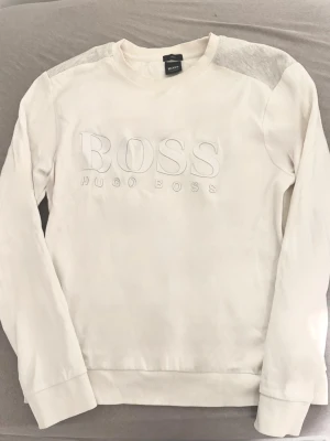 Vit sweatshirt från Hugo Boss - Vit sweatshirt från Hugo Boss med broderad logotyp på bröstet. Slim fit-modell med ribbade muddar och detaljer på axlarna. Perfekt för dig som gillar exklusiva plagg. Materialet är mjukt och bekvämt, passar till många olika outfits. det är storlek L på den men den är ju slim fit så den passar även till M