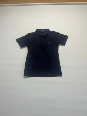 Marinblå pikétröja från Ralph Lauren storlek S - Klassisk marinblå pikétröja från Ralph Lauren med korta ärmar och röd broderad logga på bröstet. Tröjan har krage och knäppning framtill, perfekt för en stilren look. Tillverkad i mjuk bomull för skön komfort. Tröjan har storlek S och är helt ny och aldrig buren