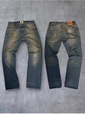 Levi's 501 - Klassiska blå jeans från Levi's med rak passform och fem fickor. Jeansen har slitningar och ljusare partier framtill och baktill för en cool, vintage look. Tillverkade i denim med detaljerade sömmar och ikonisk läderpatch bak i midjan.