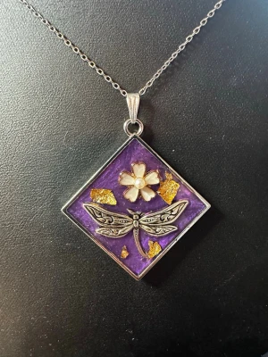 Lila halsband med trollslända och blomma - Unikt halsband med fyrkantigt hänge i lila bakgrund, dekorerat med en silverfärgad trollslända och en blomma med pärldetalj i mitten. Hänget har även inslag av guldfärgade flakes och sitter på en tunn silverfärgad kedja. Perfekt för dig som gillar naturinspirerade smycken.