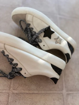 Vita sneakers med stjärndetaljer - Snygga vita sneakers med svart stjärna på sidan och glittrig silverstjärna. Skorna har svartglittriga detaljer på hälen och mönstrade svarta/vita skosnören. Ovandelen är i läder och tån är i beige mocka. Perfekta för dig som gillar unika detaljer.