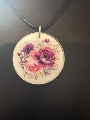 Halsband med lila blommor på rund bricka - Unikt halsband med en rund, vit bricka som har lila och rosa blommor. Hängs i ett svart snöre och passar perfekt för dig som gillar romantiska och konstnärliga accessoarer. Materialet är troligtvis keramik eller porslin.