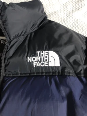Blå dunjacka från The North Face - Snygg blå dunjacka från The North Face med svarta axelpartier och broderad logga på bröstet. Jackan har hög krage, dragkedja framtill och två sidofickor med dragkedja. Perfekt för kalla vinterdagar och riktigt skön att bära.