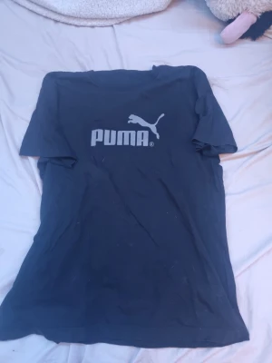 Svart Puma t-shirt med logga - Snygg svart t-shirt från Puma med stor vit logga och text på bröstet. Klassisk passform och rund halsringning. Perfekt till jeans eller shorts för en sportig look.