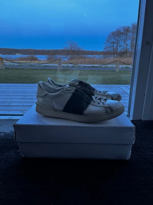 Valentino skor  - Snygga vita sneakers från Valentino med en bred svart läderdetalj över sidan. Klassisk låg modell med rund tå och vita skosnören. Skorna har en stilren design och är tillverkade i läder för en exklusiv känsla. Storlek 44, skick 6/10 använda men har mycket kvar, allt og hänger med. Pris går att diskutera✅